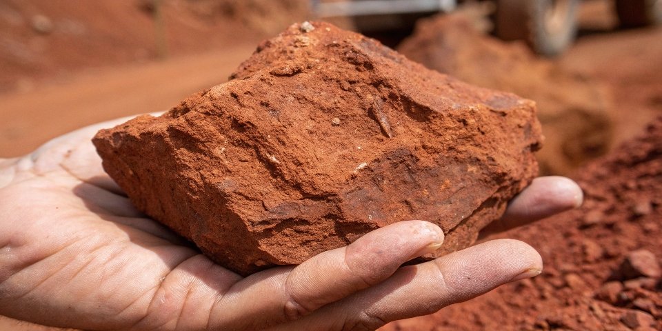 A close-up photograph of a hand holding a raw, reddish-brown bauxite rock, showing its texture.