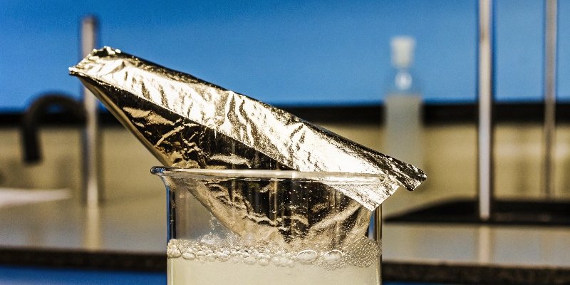 Dissolving Aluminum A piece of shiny aluminum foil reacting and fizzing in a beaker of clear chemical solution.