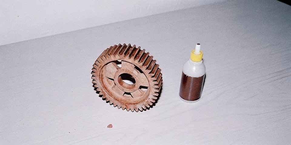 A severely pitted and damaged aluminum gear next to a bottle labeled with a caustic warning symbol