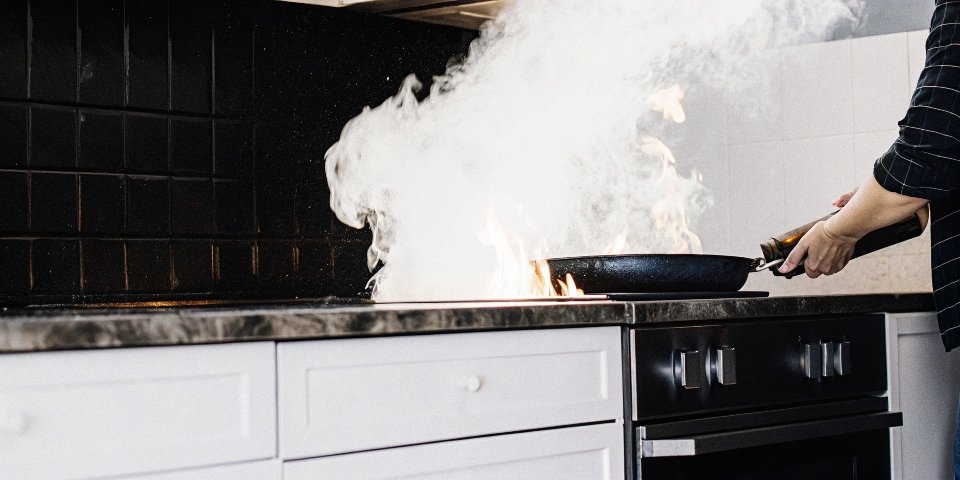 A kitchen grease fire in a pan erupting as someone tries to pour water on it