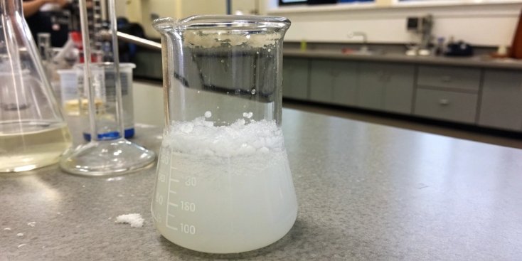 A beaker showing the precipitation of white aluminum hydroxide gel from a clear solution.